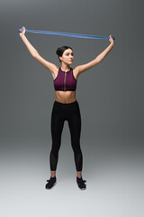 full length view of slim sportswoman training with resistance band on grey background.
