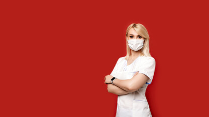 Confident female doctor wearing protective face mask standing on red background. Young woman intern in medical clothes posing.