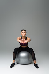 Obraz premium pretty sportswoman stretching while sitting on fitness ball on grey background.