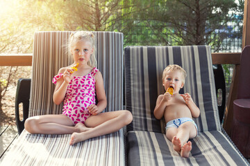 Fototapeta premium Two cute adorable caucasian blond little siblings enjoy having fun relaxing and eat fruit popsicle icecream sitting on sunbed in yard garden near pool sea beach. Brother and sister chilling outdoor
