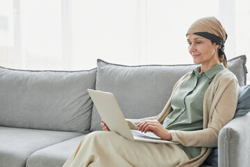 Candid portrait of mature woman using headscarf and using laptop while relaxing on couch in cozy home setting, copy space - obrazy, fototapety, plakaty