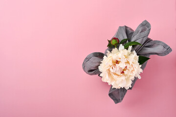 Spring eco-friendly gift wrapped in grey textile with one white peony flowers decor on pink. Sustainable gift. Traditional Japanese Furoshiki style. View from above.