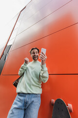 low angle view of amazed man in sweatshirt holding smartphone near orange wall.