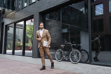 full length of happy businessman in earphones walking and holding laptop near building and bicycle.