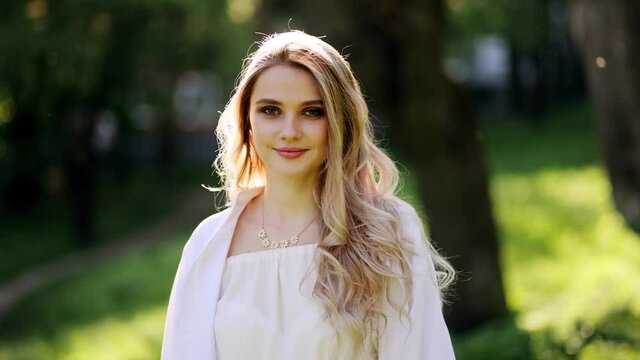 Beautiful Smiling Young Blonde Woman with Clean and pefect Skin relaxing in the park. Happy girl walking in the park. Portrait of cheerful university student looking at camera while adjusting hair