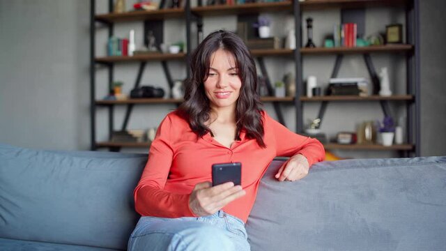 Beautiful Middle Eastern woman in red text messaging on cell phone sitting on couch at home. Female smiling and feeling surprised while reading sms and texting back on mobile phone