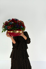 Young woman in black dress standing on white background holding red color floral arrangement in her hands. Valentine flowers. Red roses blue carnation red ranunculus bouquet in red ceramic vase.