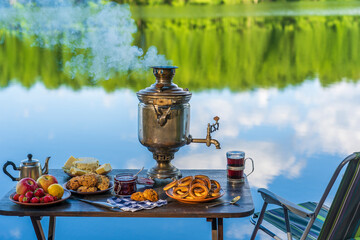Vintage metal tea samovar with white smoke and food on the table near the calm water lake in green...