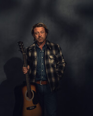 Man in checkered jacket with acoustic western guitar in dappled sunlight in front of a grey wall.
