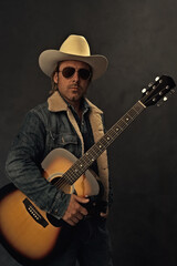 Man in denim jacket, white cowboy hat and aviator sunglasses with acoustic western guitar in front of a grey wall.