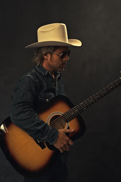 Man in denim shirt, white cowboy hat and aviator sunglasses with acoustic western guitar in front of a grey wall.