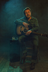 Obraz premium Musician sitting on a wooden stool with an acoustic guitar with next to him a guitar case with a glass of whiskey and an ashtray.