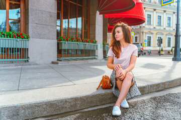 A young woman in a skirt sits on the sidewalk under the red awning of the famous Astoria Hotel in...