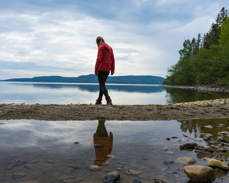 Girl wearing red jacket walking on the beach by a lake in the High Coast area, Northern Sweden. Reflections in the water, cloudy sky.