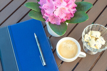 A tablet with a pen and in a vase white chocolate and an alpine rose on a wooden table and a cup of coffee with sweets,