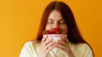 Young woman holding in hands and sniffing fresh large juicy strawberries. over yellow background