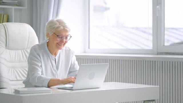Smiling aged lady with grey hair and glasses types on laptop