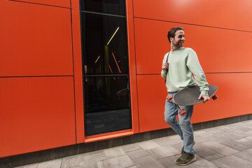 cheerful man in sweatshirt holding longboard and walking near orange wall.