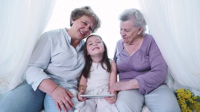 Old great-grandmother and grandma nanny kissing and hugging cute small preschool little girl with digital tablet. Three generation family at home, Female family enjoy, respect and warm relation