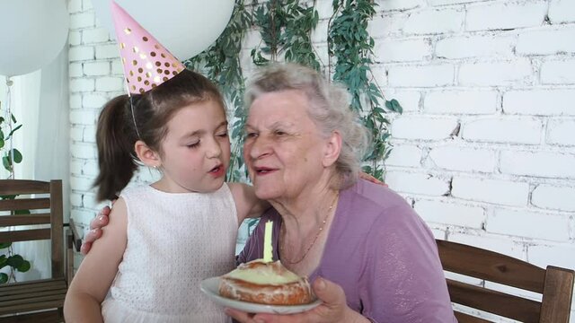 Happy smiling little cute girl hugs and kisses her senior grandmother on celebration 85th birthday, old lady holds cake with extinguished candle. Enjoy, respect and warm relation. Celebration concept