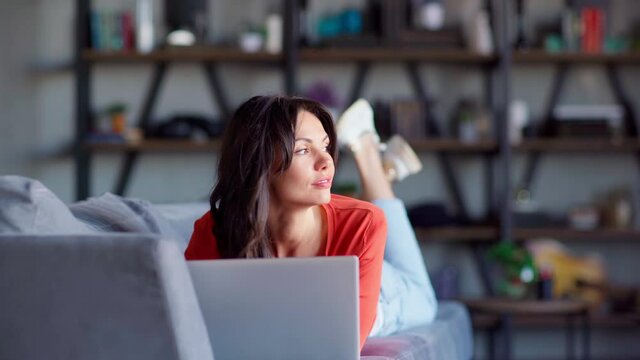 Front view of attractive middle aged brunette woman lying on sofa at home, using laptop computer (working or browsing social media) and thinking