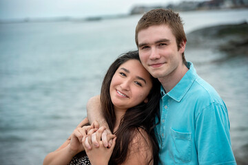 Obraz premium Portrait of young smiling happy couple in romantic casual loving embrace on a warm summer day at the beach with blue sky and water in background.