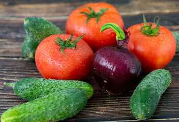 Simple organic composition on the wooden background. Fresh tomatoes, cucumbers and red onion with bright greens. Tasty summer vegetables with water drops. 