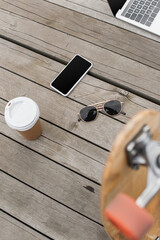 high angle view of gadgets with blank screen, sunglasses and paper cup on wooden table near blurred longboard.