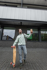 Fototapeta premium full length of joyful man in sweatshirt holding paper cup and longboard outside.