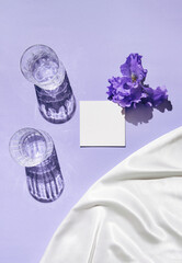 Two glasse with water and card note with iris flower on pastel lilac background with white silk cloth. Summer refreshment concept. Sunlit flat lay. Minimal style. Top view