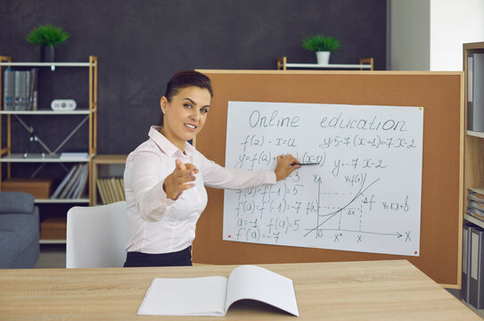 Portrait of female teacher explaining equations in online class. Beautiful young woman sitting at desk, looking at camera, pointing at classroom board and asking question during virtual math lesson