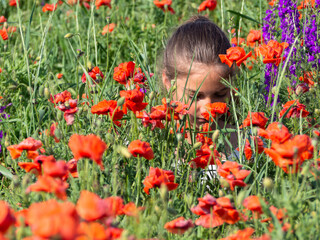 The girl sits among the red poppy flowers in the field and enjoys their smell. A teenage girl is happy and in harmony with nature. The concept of peace and love