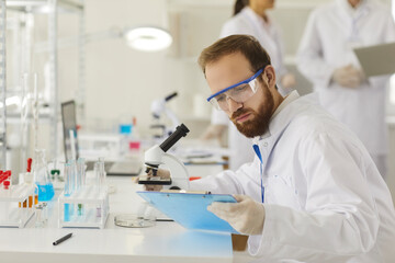 Medical male pharmacy scientist wearing protective clothes and goggles working in laboratory comparing test sample reading paper report on clipboard sitting at desk. Medicine and biotech