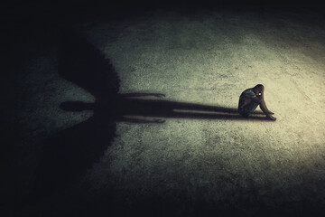 Depressed young man sitting alone isolated in a dark room, casting a shadow of a powerful guardian angel. Inner strength conceptual background. Stressed person introvert with an imaginary friend