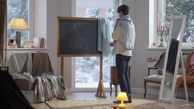 Wide shot of talented fashion designer drawing dress sketch on blackboard with chalk. Inspired Caucasian young man creating design indoors in atelier studio. Talent and creativity