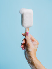 Woman's hand holding a wooden stick with a white soap with foam on top isolated on a bright blue background. Abstract and surreal pop art. Creative summer hygiene or dessert concept.