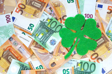 Green felt four leaf clover on surface covered with 50 and 100 euro banknotes. Financial, bank,...