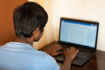 Portrait of a young Indian teen typing on laptop. Indian student uses laptop to complete his assignment. Online education concept. Kid typing on his laptop to complete his school project.	