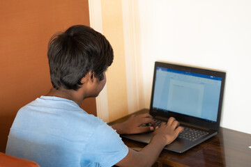 Portrait of a young Indian teen typing on laptop. Indian student uses laptop to complete his assignment. Online education concept. Kid typing on his laptop to complete his school project.	