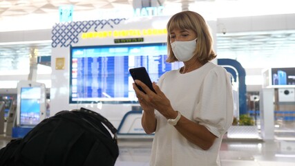 Middle aged woman wearing face mask checking her phone standing by the departure board at the airport 