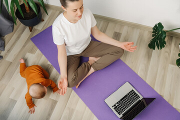 Selective focus of calm mom watching yoga tutorial online on laptop doing yoga sit on warm floor at home, caucasian mother meditating for stress relief relaxing in living room with cute baby daughter