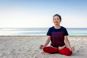Senior Asian woman doing yoga Padmasana sitting pose (lotus pose)  for meditation on sandy beach with summer blue sky and sea. Healthy elder people, relax and calm concept. Phuket, Thailand.
