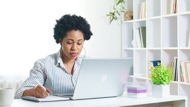 African American female student uses a laptop and writes in a notebook. Portrait of a young black woman working remotely on the computer at home. Online education 4K.