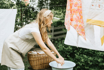 blonde teen girl in linen apron is doing homework washing clothes in basin and hanging them on...