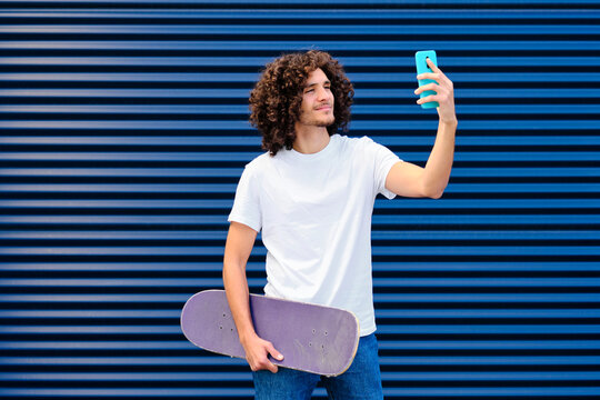 Smiling young man with skateboard taking selfie through smart phone