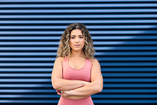 Young confident female sportsperson standing with arms crossed in front of blue wall