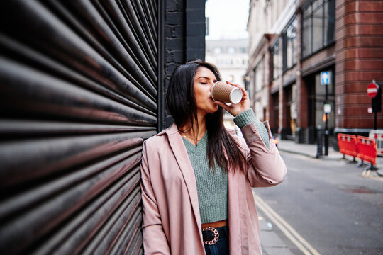 Beautiful woman in overcoat drinking coffee while standing by closed shutter in city
