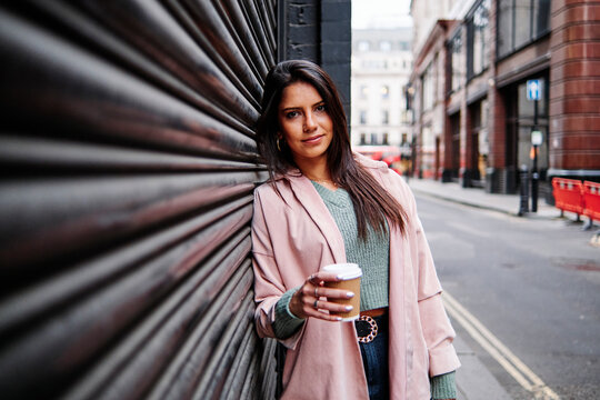 Smiling woman with coffee cup leaning on shutter in city