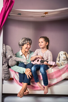 Granddaughter playing guitar while sitting by grandmother on bed