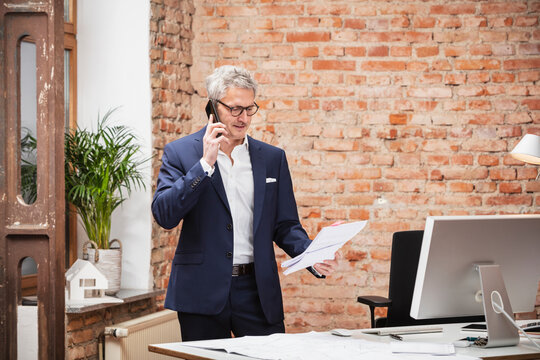 Mature male architect with document talking through smart phone by desk in office